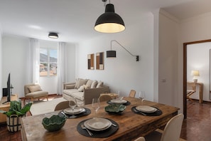 A modern living room with a wooden dining table set for four, a sofa, a TV, and a view of the city.