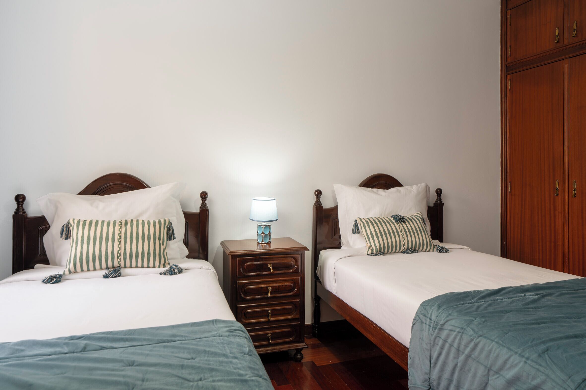 A room with two single beds, each with a wooden headboard and white bedding. There is a nightstand with a lamp between the beds.