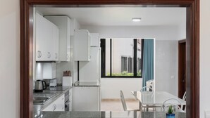 Une cuisine moderne avec des armoires blanches, un évier en acier inoxydable et une machine à café. On y trouve un coin repas avec une table et des chaises blanches, ainsi qu’une fenêtre donnant sur l’extérieur.