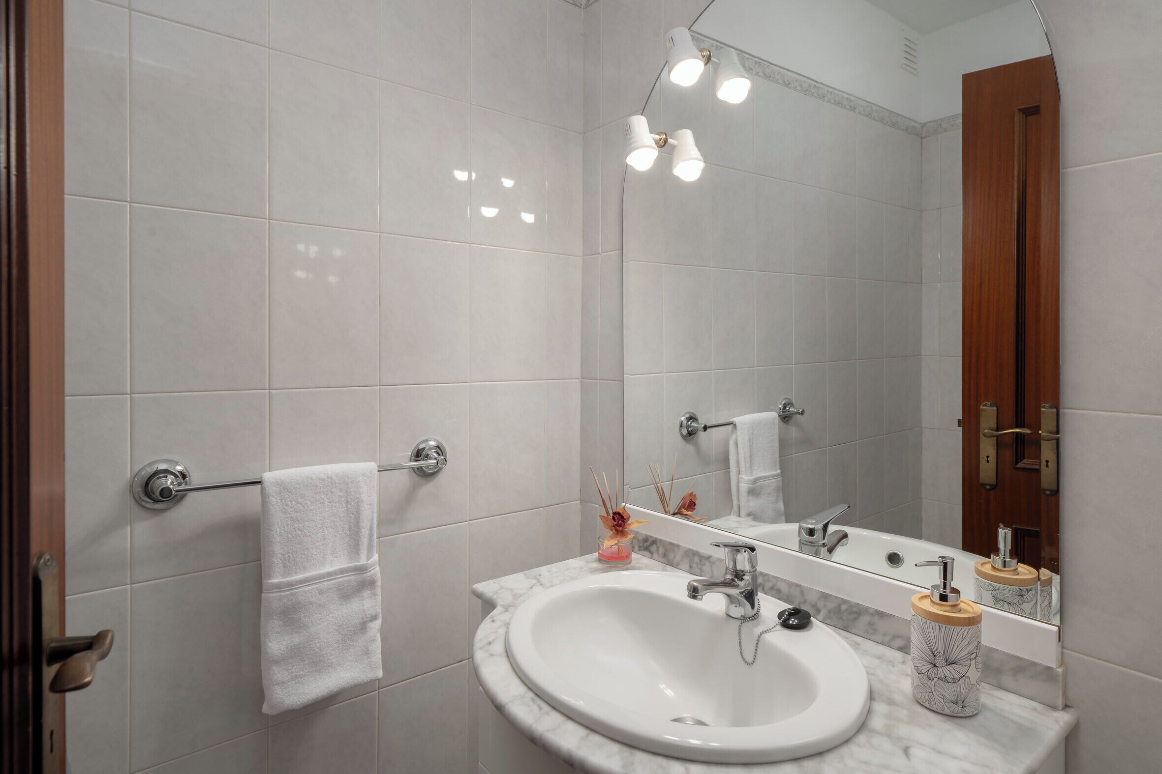 Casa de banho com azulejos brancos, lavatório branco, espelho e porta-toalhas.