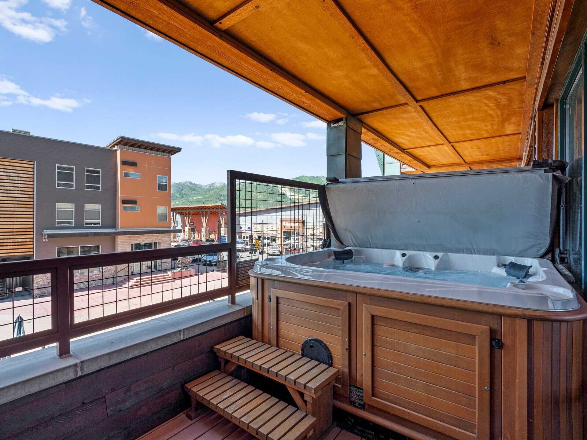 Un jacuzzi en un balcón con barandas de madera, vista a edificios y cielo despejado.