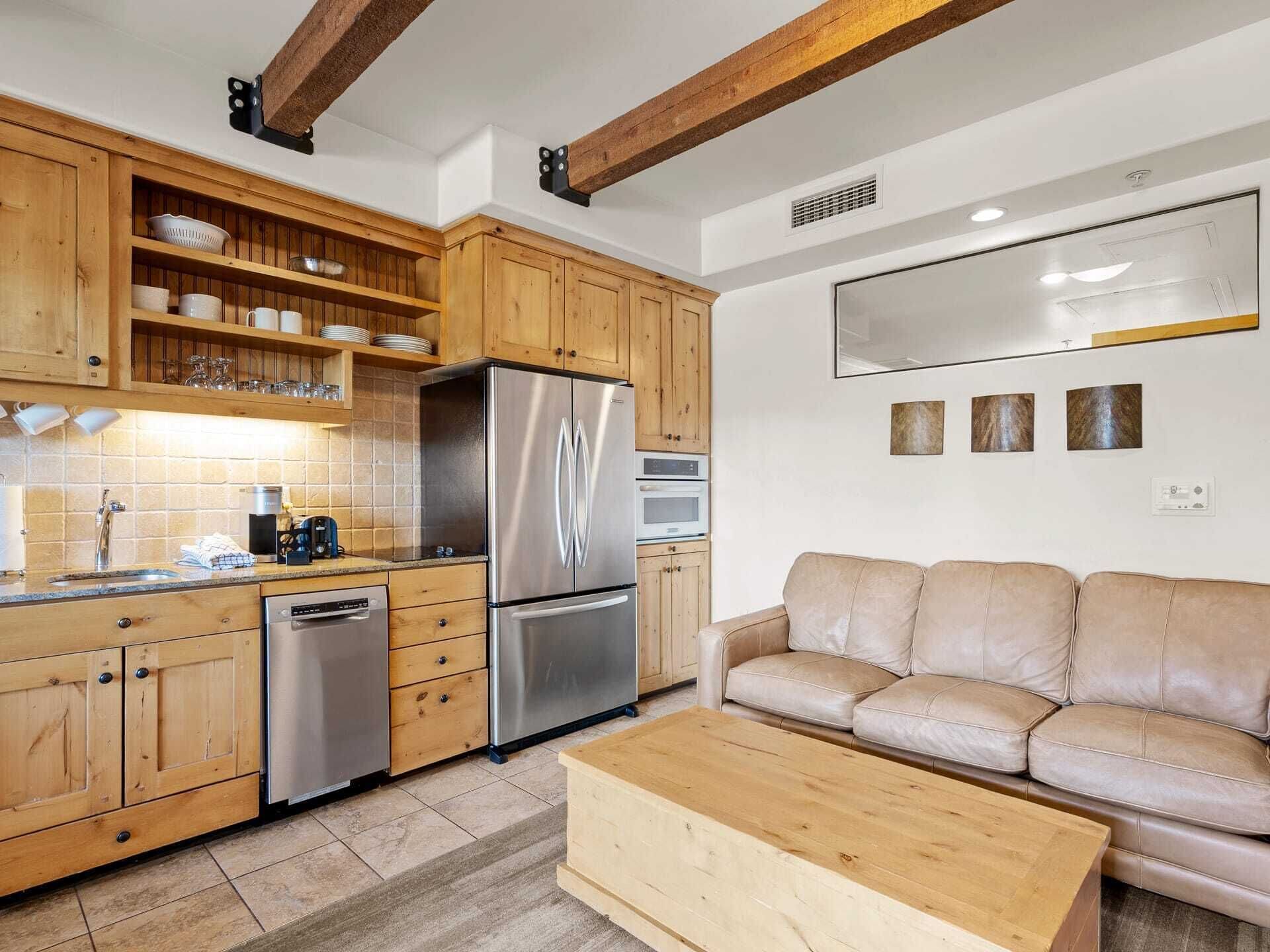 A modern kitchen with wooden cabinets, a stainless steel refrigerator, and a beige leather sofa.
