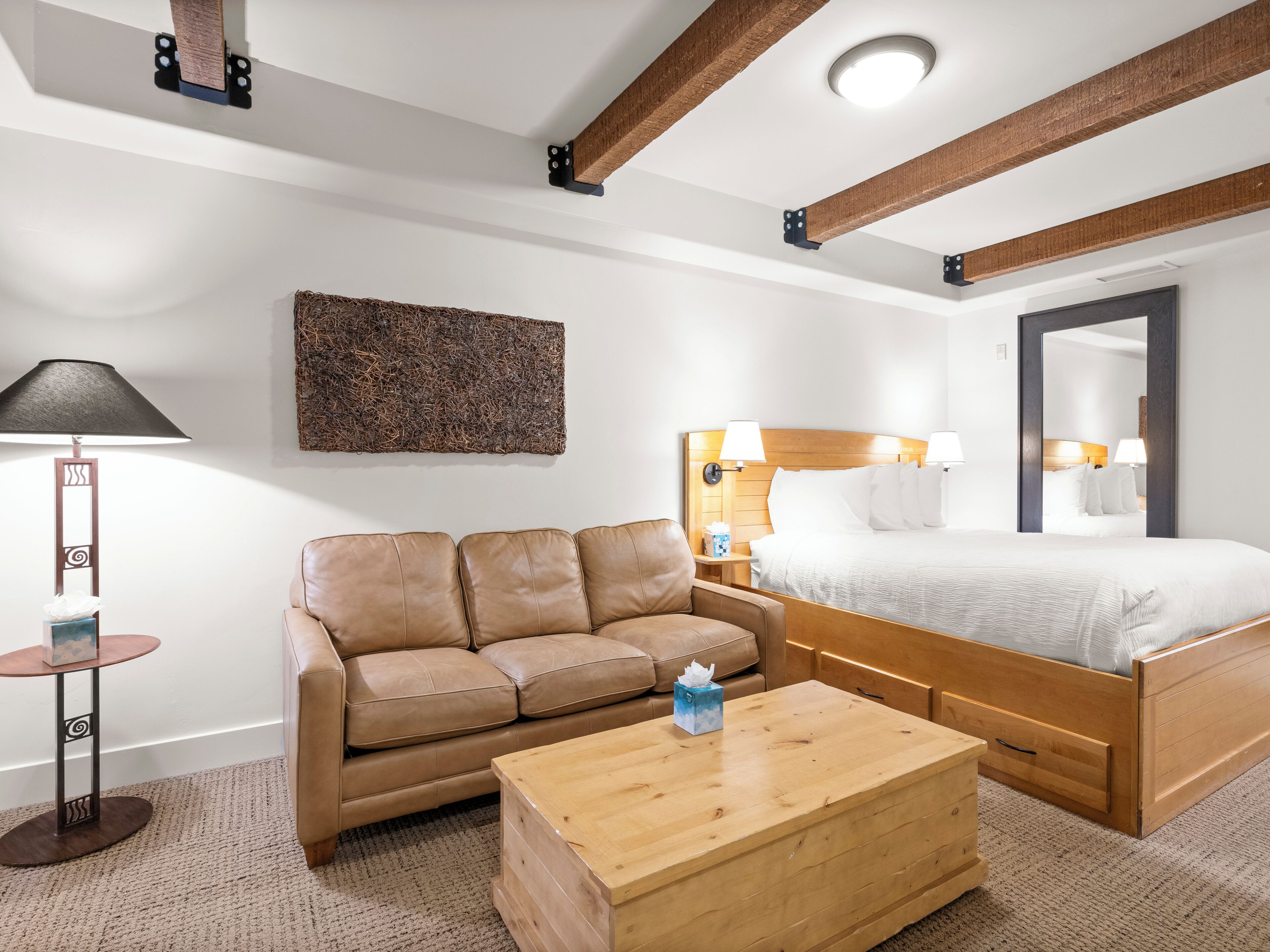A hotel room with a wooden bed, a leather sofa, a wooden coffee table, a wall-mounted lamp, and a textured artwork.
