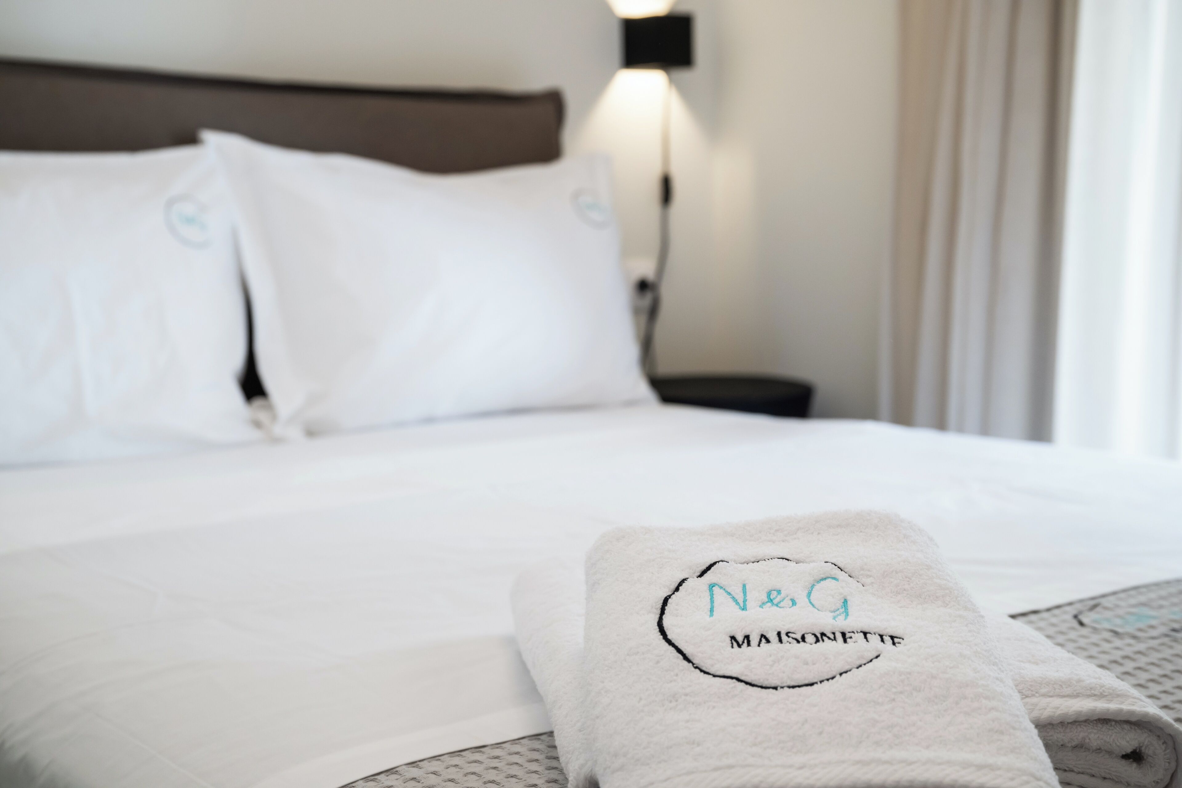 A neatly made bed with white linens and a folded towel with "Neg Maisonette" embroidered on it.