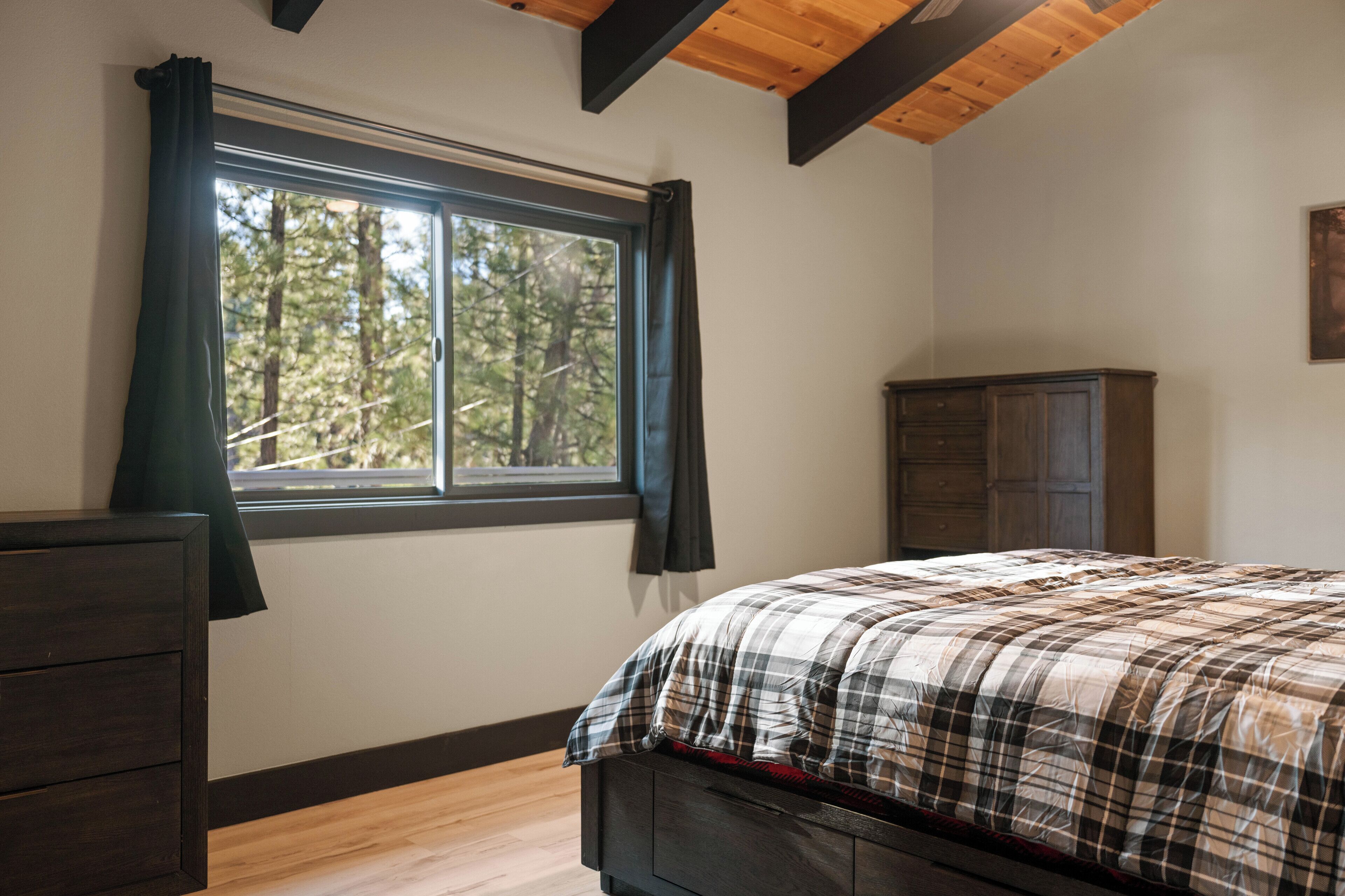 A bedroom with a large window, a bed with a plaid bedspread, a dark wooden dresser, and a wooden cabinet.