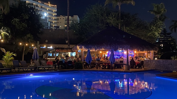 Et poolområde om natten med liggestole, en bar og en tilbygning med stråtak.