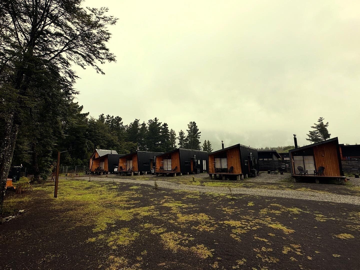 A row of wooden cabins with a gravel path in front, surrounded by trees.