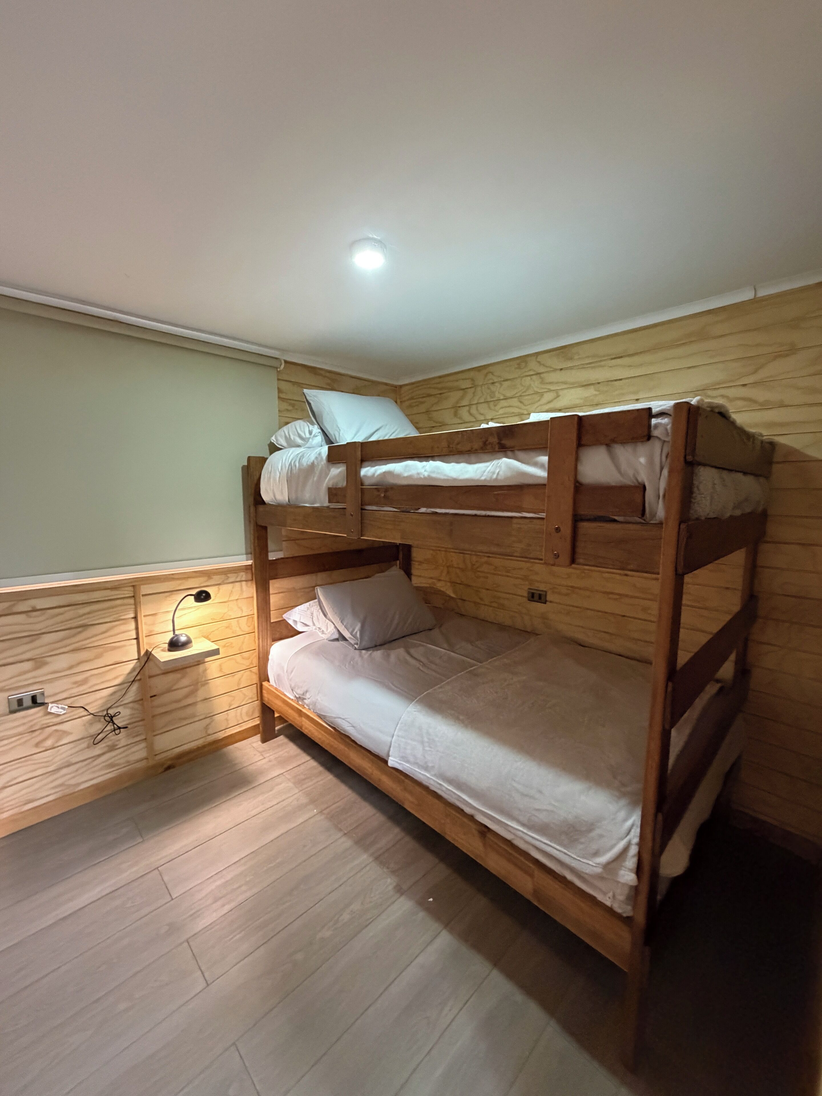 Uma cama de beliche com cabeceira de madeira, mesa de cabeceira com abajur e uma tomada na parede.