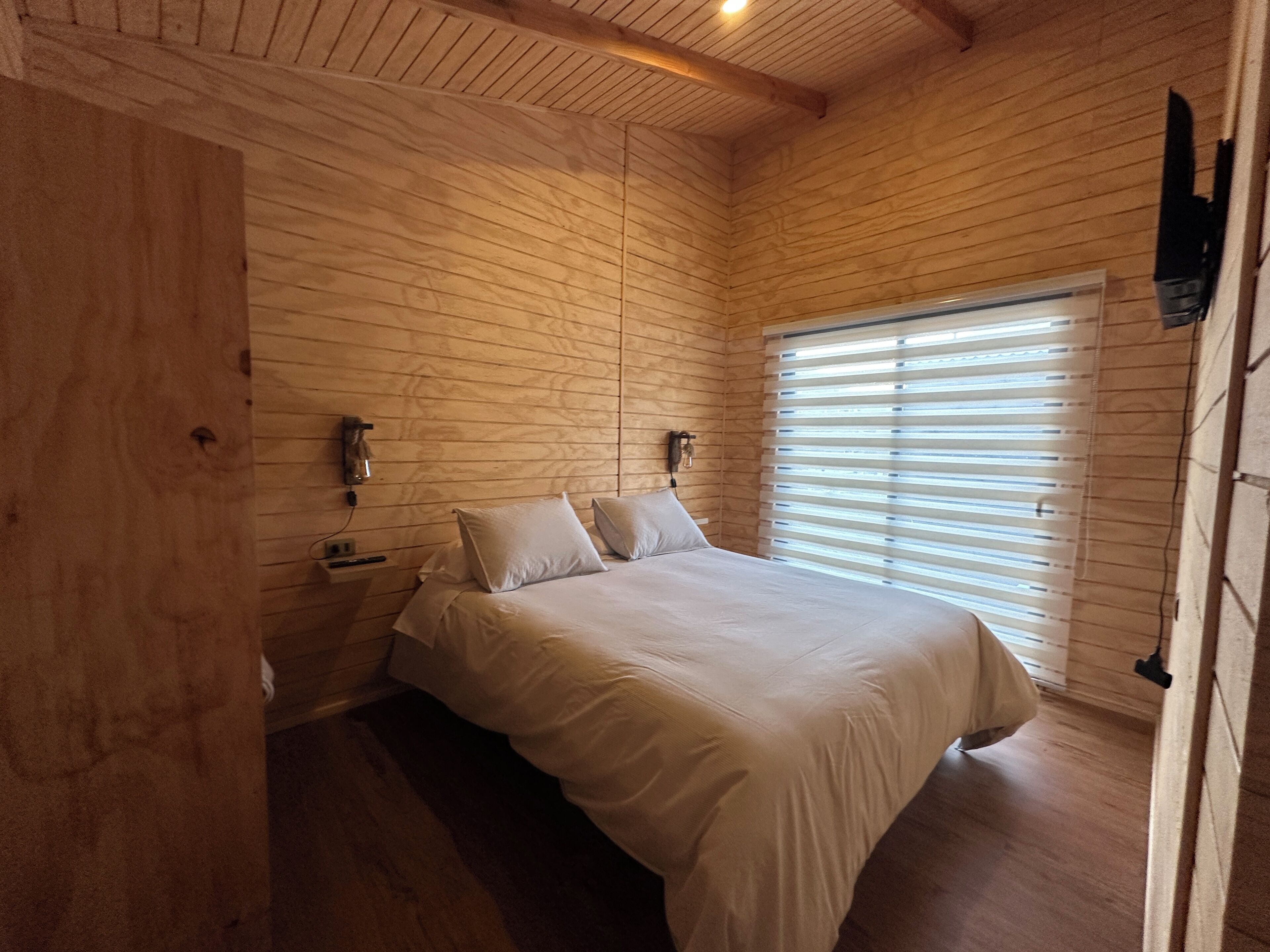 Een slaapkamer met houten lambrisering, een bed, nachtkastjes en een raam met zonwering.