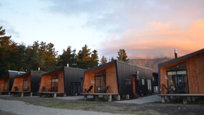 Une rangée de chalets modernes aux grandes fenêtres et aux façades en bois, se dressant sur un fond de forêt et d’un ciel nuageux.