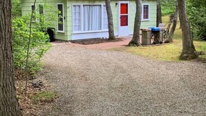 A small, single-story house with a green exterior and a red door, surrounded by trees and a gravel driveway.