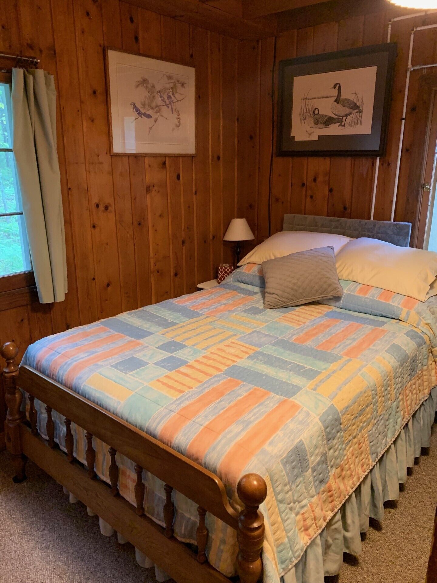 Quarto com painéis de madeira, uma cama com colcha colorida, mesa de cabeceira com abajur e quadros nas paredes.