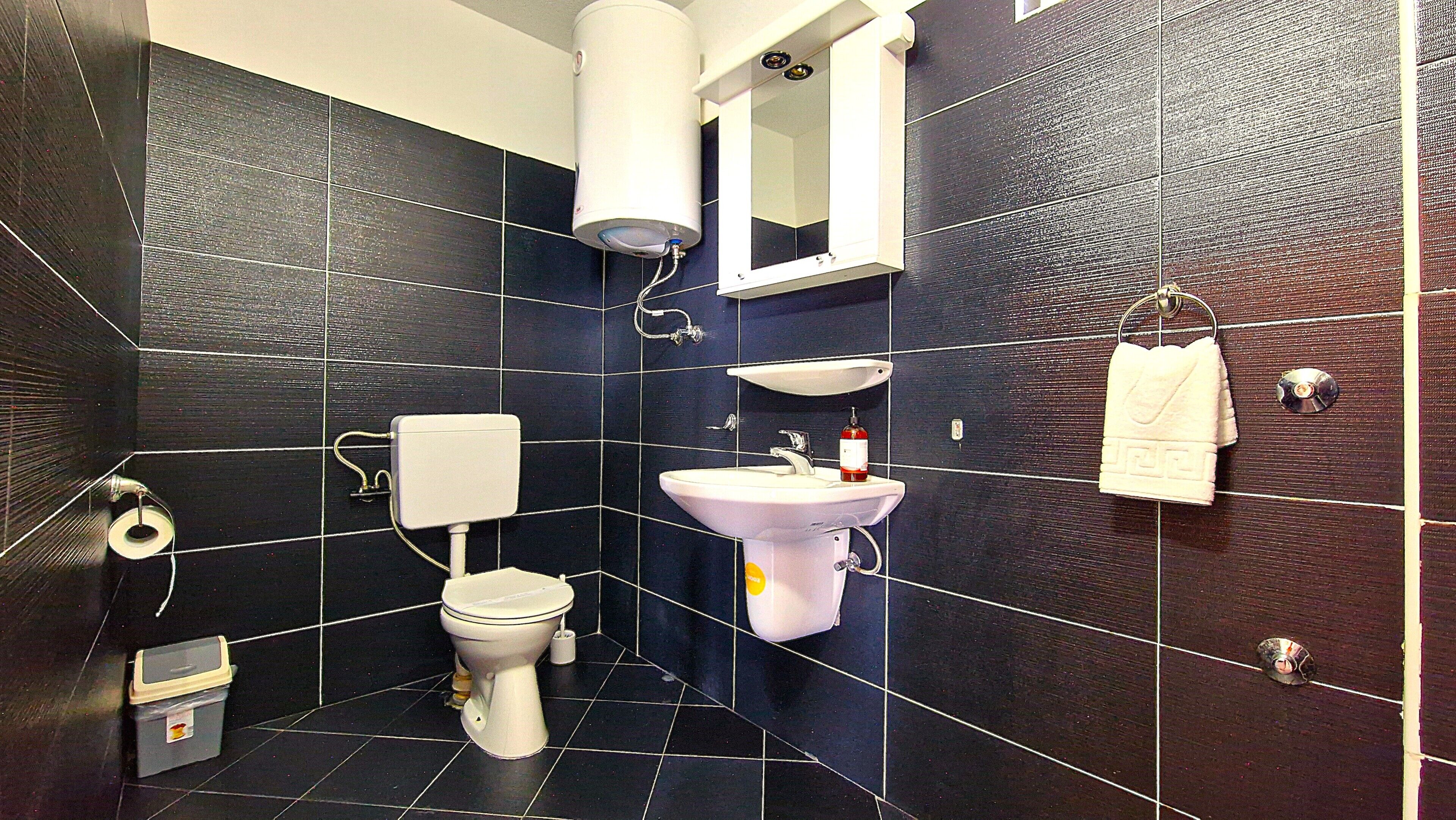 Egy fürdőszoba, amelyben található egy WC, mosdókagyló, tükör, törölközőtartó és WC-papírtartó.