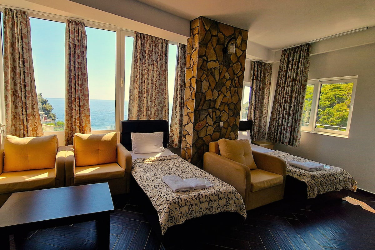Habitación de hotel con sofá, dos camas individuales, una mesita y ventanas amplias con cortinas.