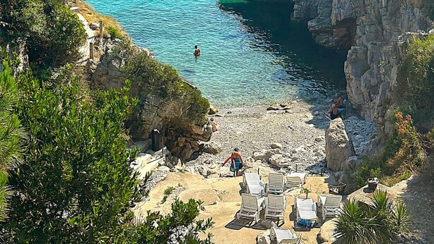 A coastal beach with clear blue waters, rocky cliffs, and a row of sun loungers. - AZURA BAY HOTEL (Dobra Voda)