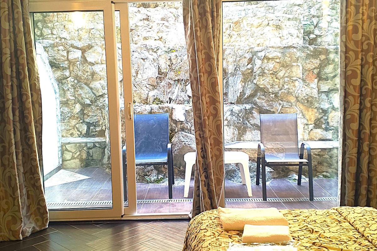 Habitación de hotel con cama, cortinas y vista a un espacio exterior con sillas y un muro de piedra.