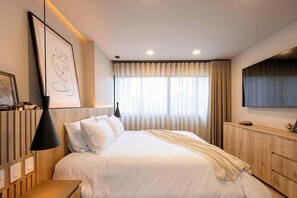 A modern hotel room with a large bed, a bedside table, a lamp, a framed artwork, a window with curtains, and a TV mounted on the wall.