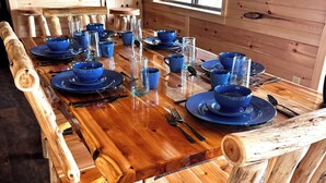 Una mesa de comedor de madera con platos y vasos azules, rodeada de sillas de madera en una habitación tipo cabaña con paredes de madera y una ventana.