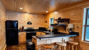 Cocina compacta con paredes de madera, un refrigerador negro, estufa y pileta. Hay dos taburetes de madera y un pequeño estante con vajilla.