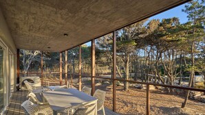 Un espace extérieur abrité, doté d’une table et de chaises, de grandes fenêtres et offrant une vue sur les arbres et le paysage.