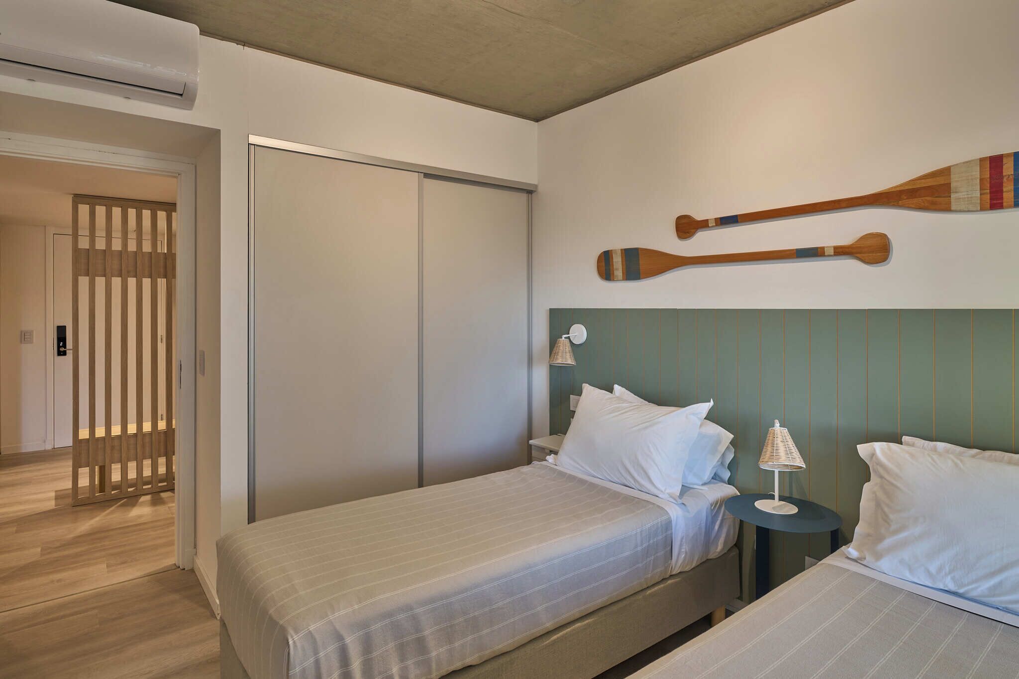 A hotel room with two beds, a wardrobe, a wall-mounted paddleboard, and a small table with a lamp.