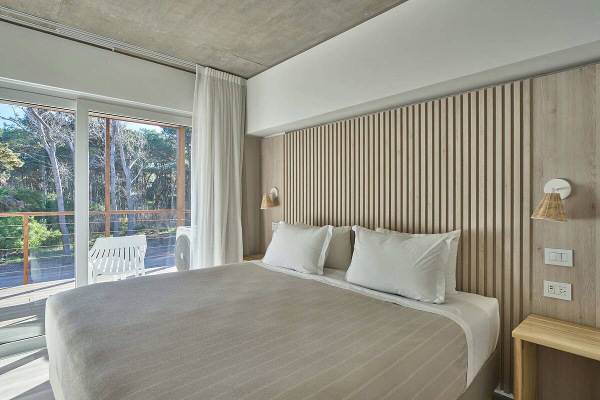 A modern bedroom with a large bed, a wooden slatted headboard, a white bench by the window, and a view of trees outside.