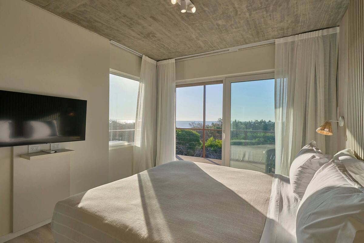 A modern bedroom with a large bed, a flat-screen TV mounted on the wall, and a balcony with a view of the outdoors.