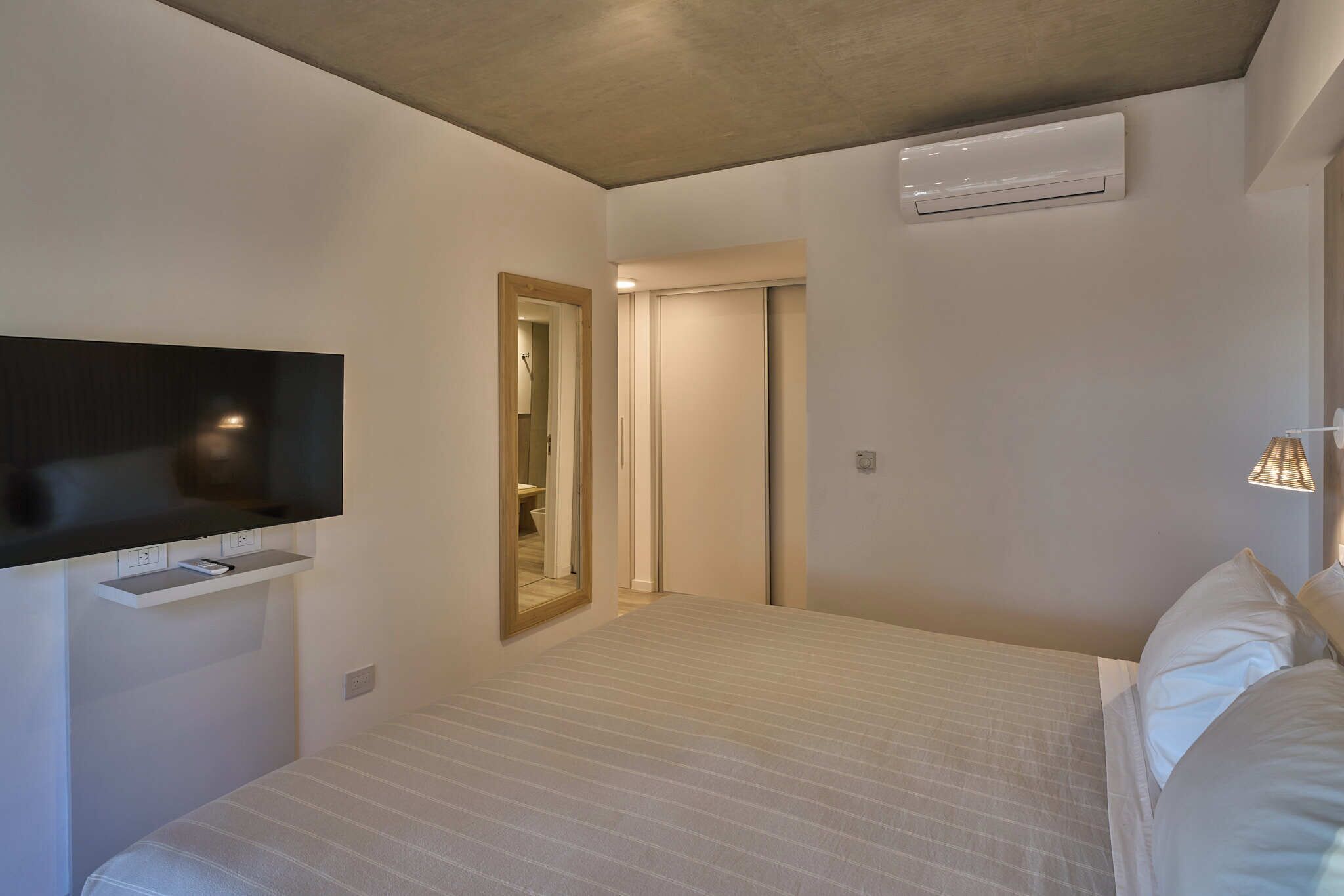 Una habitación de hotel moderna con televisión de pantalla plana montada en la pared, una cama con ropa blanca, una mesita de noche y un equipo de aire acondicionado en la pared.