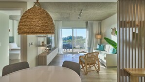 Un vaste espace de vie comprenant une grande table ronde, des chaises en osier, un canapé avec des coussins et une vue sur un balcon extérieur, accessible par une porte vitrée.