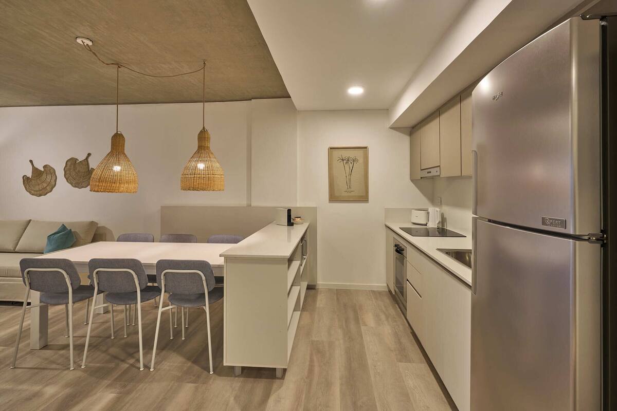 A modern kitchen with a built-in refrigerator, a dining area with a white table and chairs, and pendant lights with a woven design.