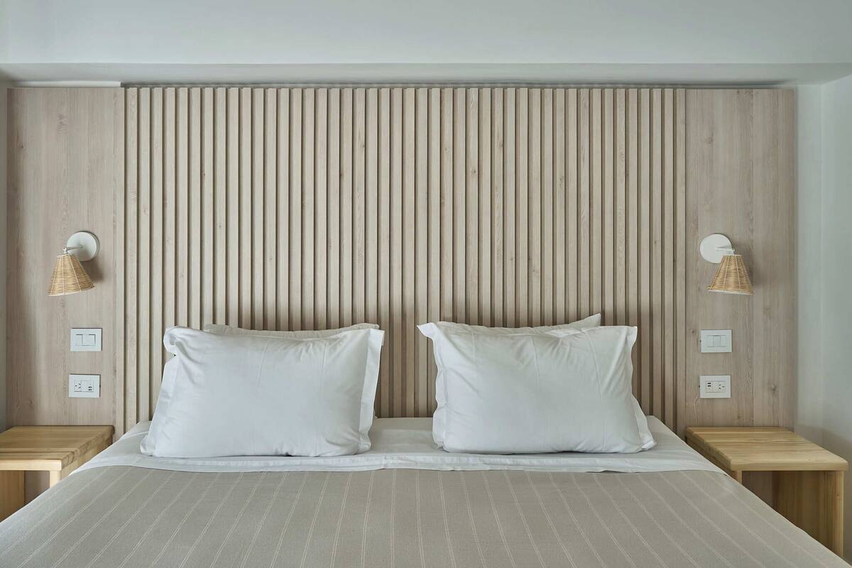 A bed with two pillows, a headboard with vertical slats, bedside lamps, and electrical outlets on the wall.