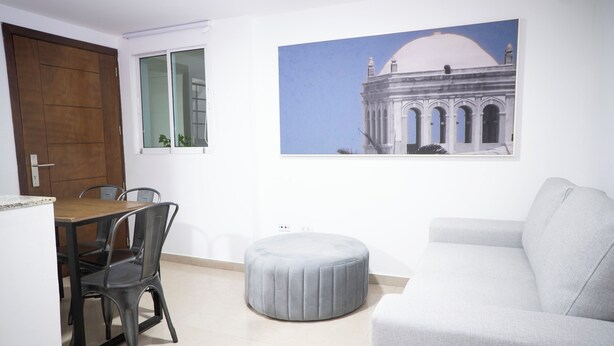 A living room with a grey sofa, a round ottoman, a dining table with chairs, and a wall-mounted picture of a building. - Apartment 5C Minaret (Bolívar)