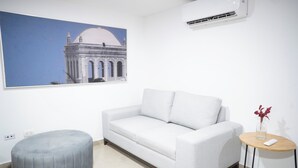 A modern living room with a grey sofa, a round ottoman, a small wooden side table with a vase, and a framed picture of a building on the wall. - Apartment 5C Minaret (Bolívar)