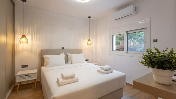 A modern hotel room with a large bed, bedside tables, a potted plant, and a window with a view of greenery. - The Secret Garden Stay (Agia Paraskevi)