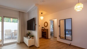 A modern living room with a flat-screen TV, wooden flooring, a console table with decorative items, a large mirror, and a sliding glass door leading to an outdoor area. - The Secret Garden Stay (Agia Paraskevi)