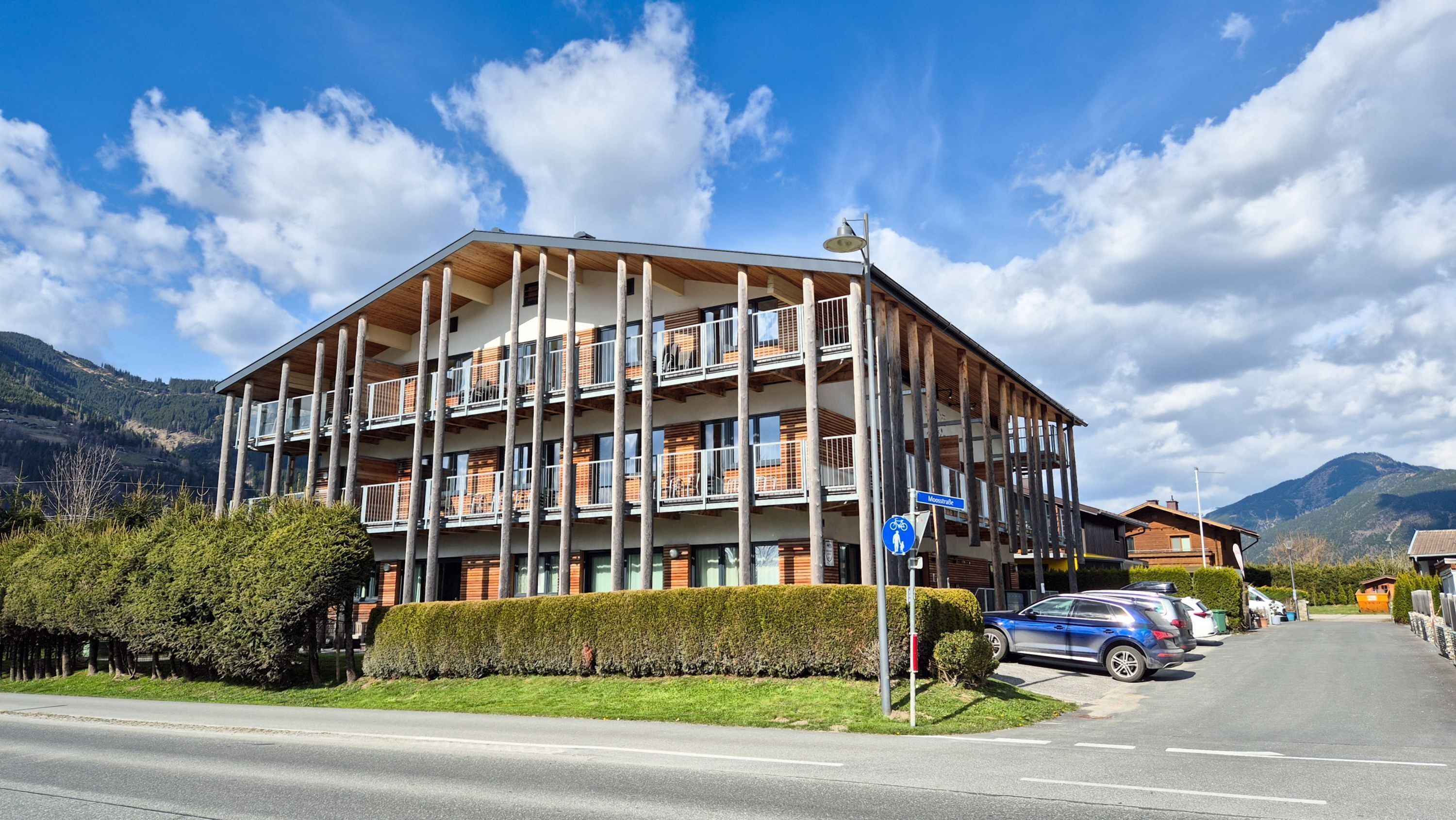 A multi-story building with balconies, a sign indicating a location, and a parking area with cars.