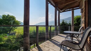 A wooden deck with a glass railing overlooking a green landscape and mountains.