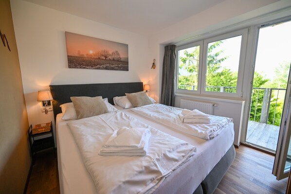 A double bed with white linens, a headboard, two bedside lamps, a nightstand, a window with curtains, and a view of greenery outside.