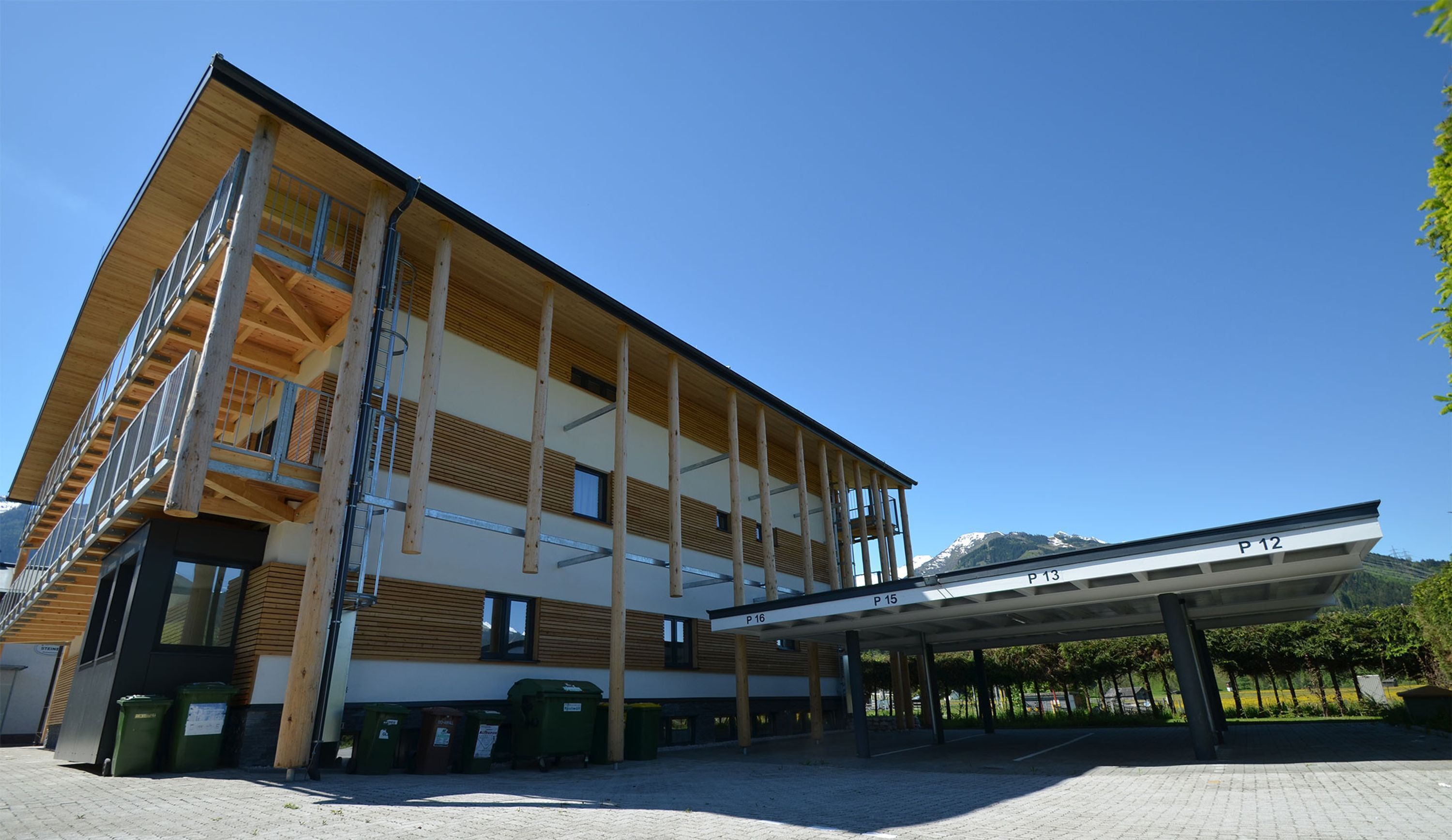 Un edificio de varios pisos con fachada de madera, una zona de estacionamiento techada y montañas al fondo.