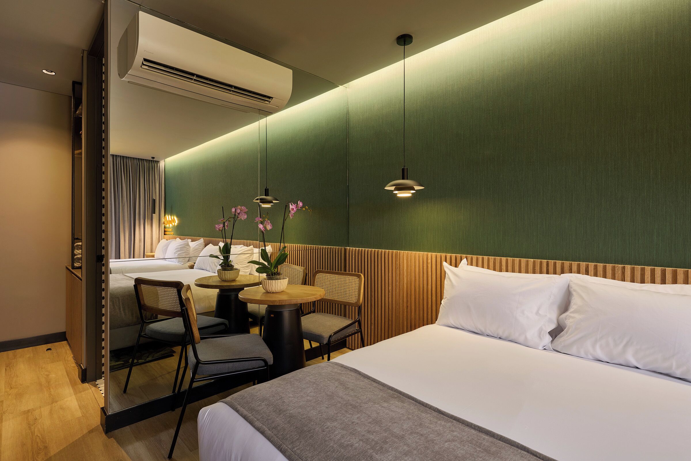 A hotel room with a bed, a desk, two chairs, a small table with a plant, and a mirror reflecting the room.
