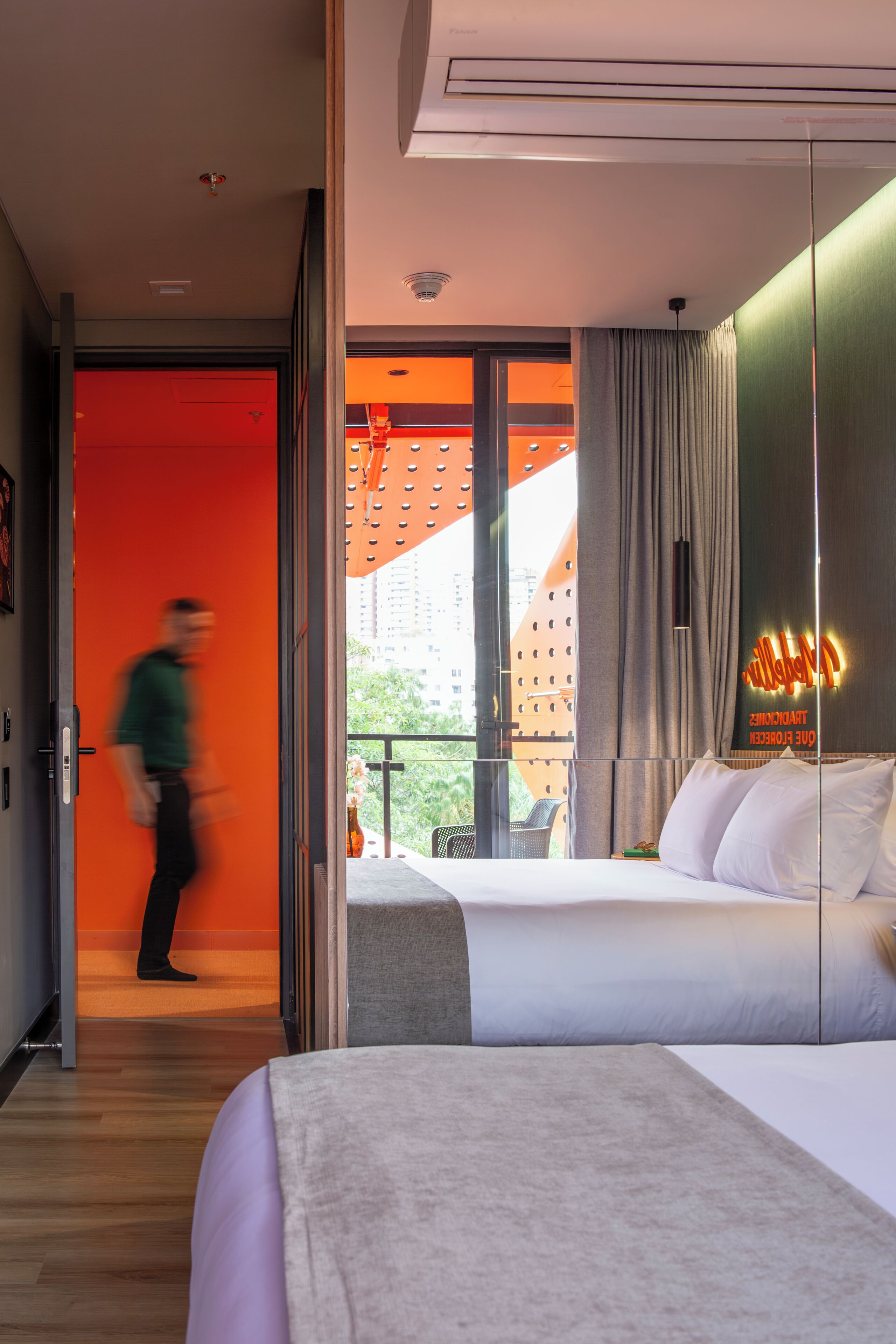 A hotel room with a bed, a nightstand, a window with curtains, and a view of buildings outside.