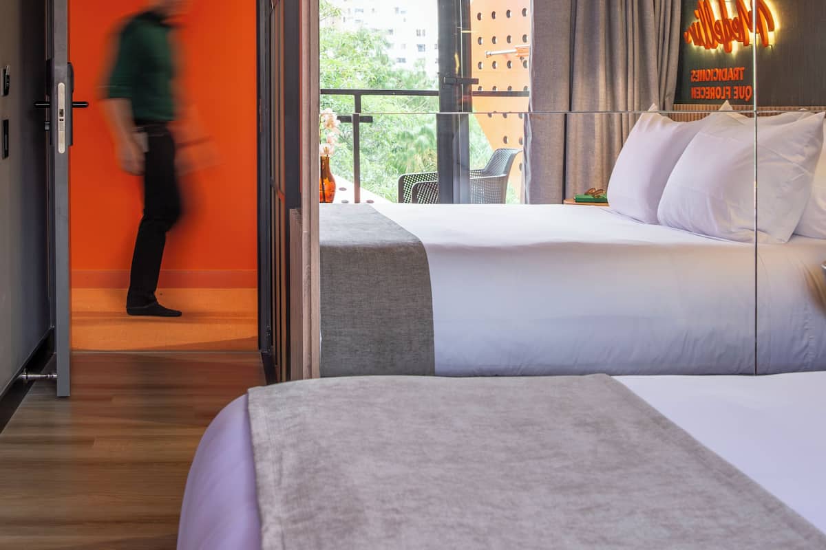 A hotel room with a bed, a nightstand, a window with curtains, and a view of buildings outside.