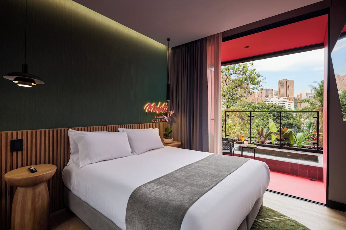 A modern hotel room with a large bed, a bedside table, a neon sign, and a balcony with a city view.
