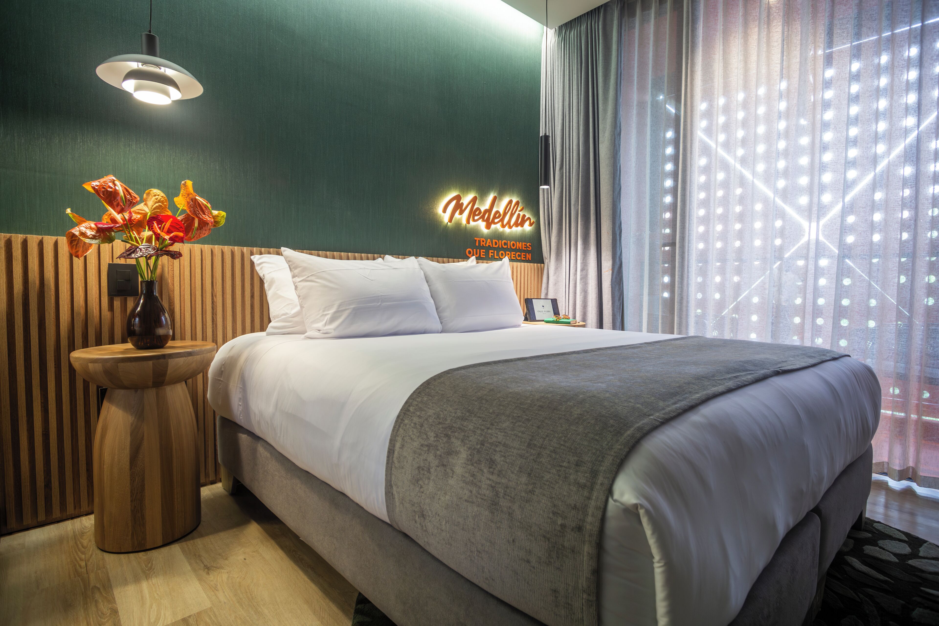 A hotel room with a large bed, a bedside table with a vase of flowers, a neon sign, and a view of the city through sheer curtains.