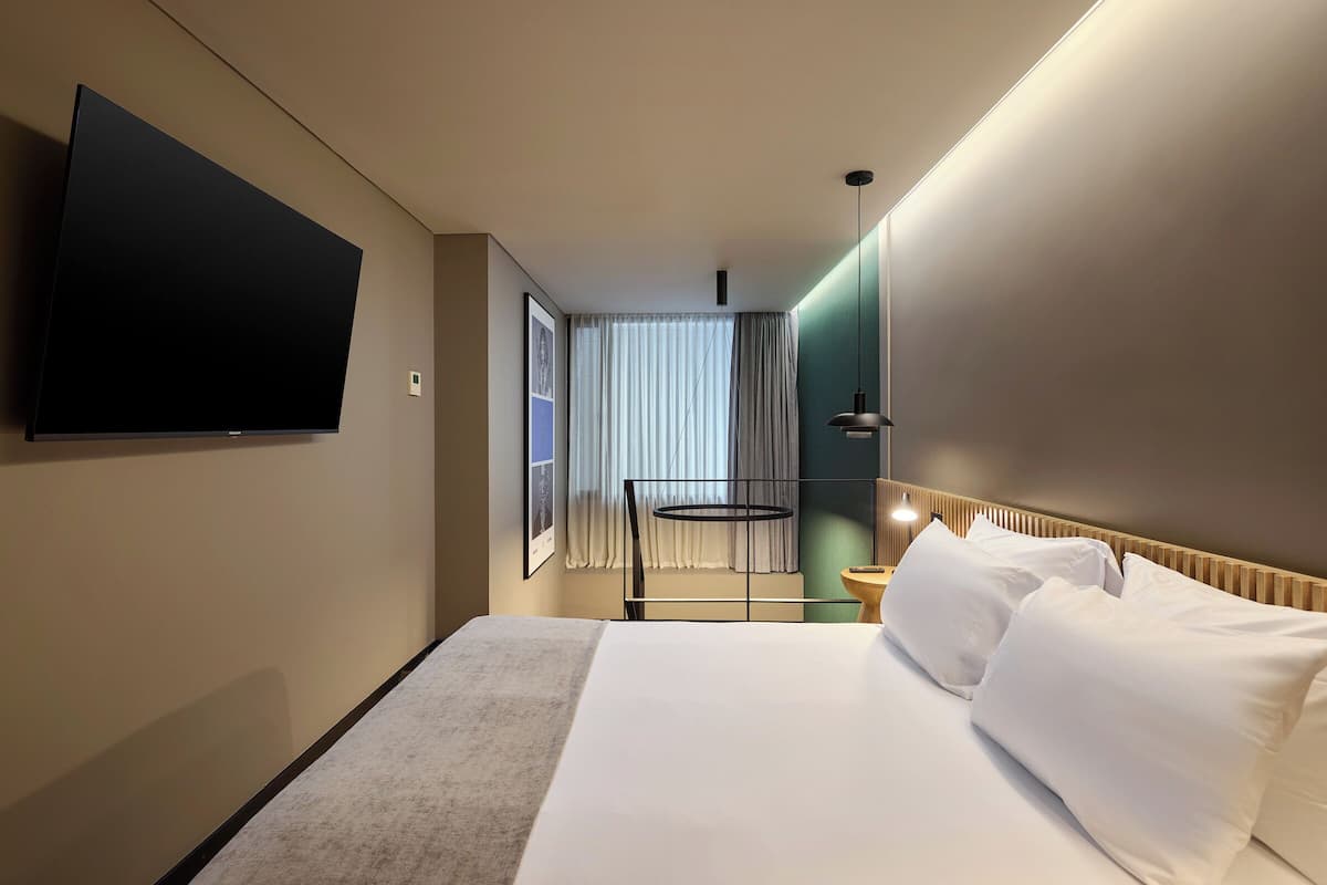A hotel room with a large bed, a flat-screen TV mounted on the wall, a window with curtains, and a bedside table with a lamp.