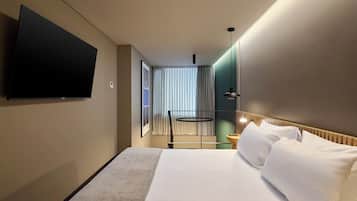 Habitación de hotel con una cama grande, un televisor de pantalla plana montado en la pared, una ventana con cortinas y una mesita de noche con lámpara.