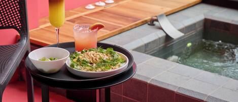 Una mesa de comedor con un tazón de ensalada y un plato de comida, un vaso de jugo, y una tabla de madera con objetos pequeños.