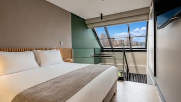 A modern hotel room with a large bed, a view of the city through a skylight, and a flat-screen TV mounted on the wall.