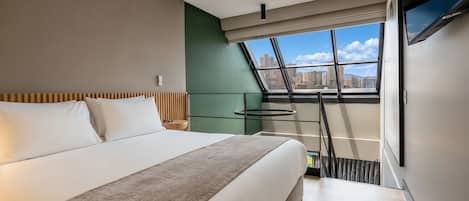 A modern hotel room with a large bed, a view of the city through a skylight, and a flat-screen TV mounted on the wall.