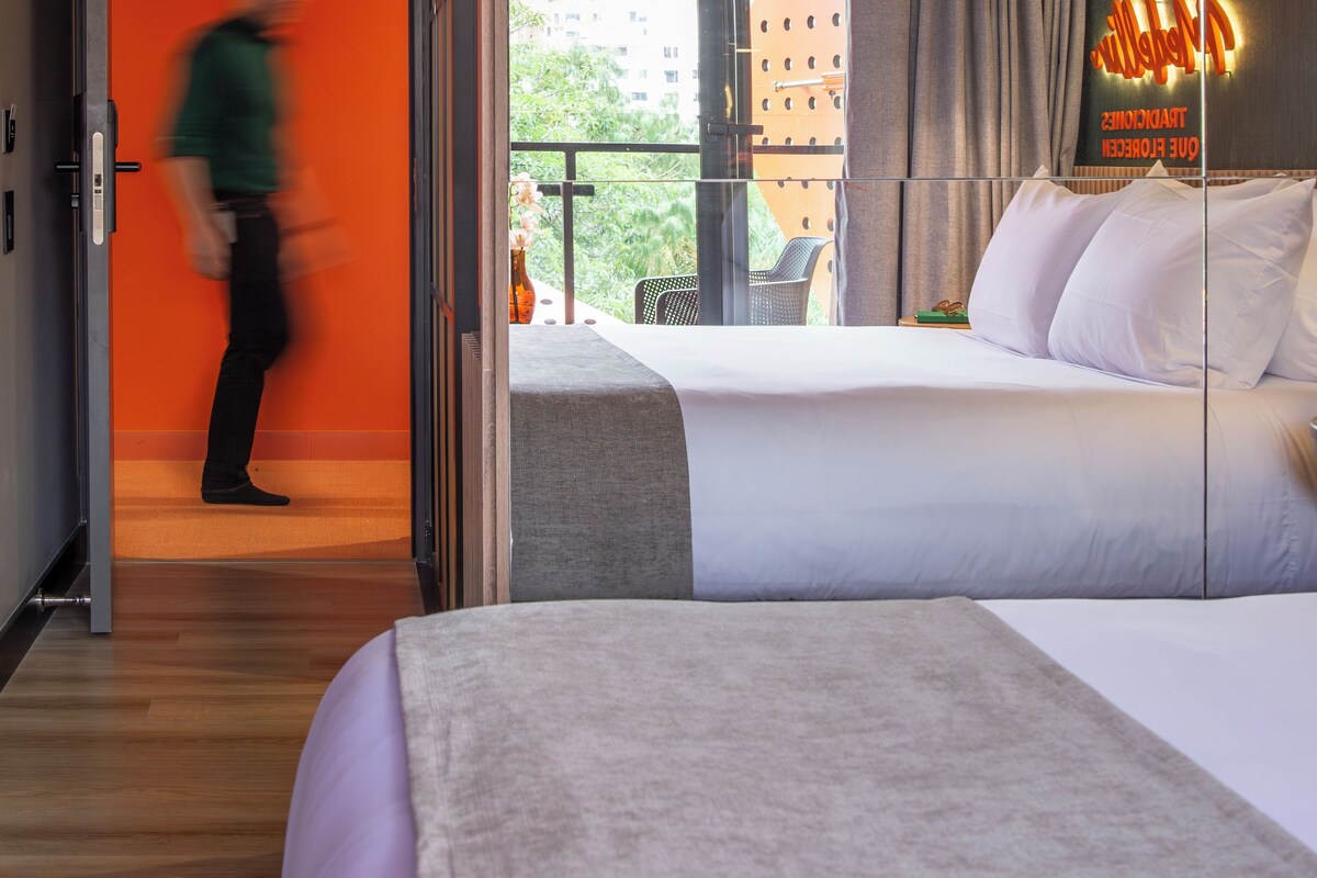 A hotel room with a bed, a nightstand, a window with curtains, and a view of buildings outside.